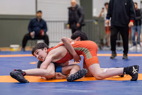 Two young wrestlers in red singlets battle on the mat, one grimacing as he's pinned down in a tight hold.