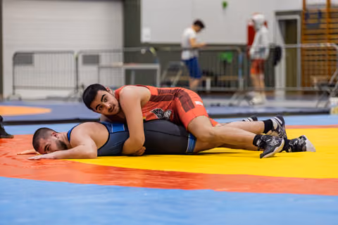 Young wrestler in red singlet pins opponent to the mat, maintaining control with focused expression in indoor sports hall.