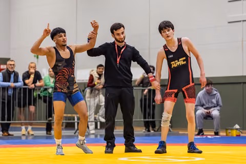 Referee raises the left arm of a jubilant wrestler in tiger singlet as the defeated opponent stands solemnly beside them.