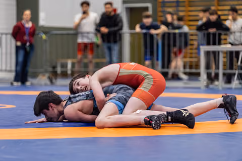 Young wrestler in red singlet dominates opponent, pinning him face-down on the orange and blue mat.
