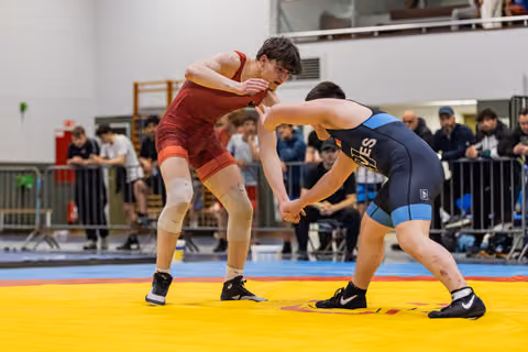 Two young male wrestlers lock grips in a tense standoff on a yellow mat, spectators watching from behind barriers.