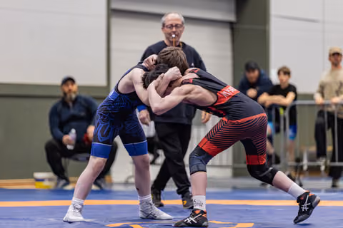 Two young wrestlers in a fierce clinch on the mat, referee watching closely behind them in an indoor sports hall.