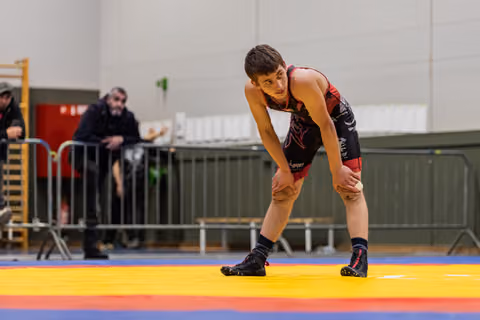Teen wrestler in black and red singlet crouches in ready stance on colorful mat, looking focused and tired.