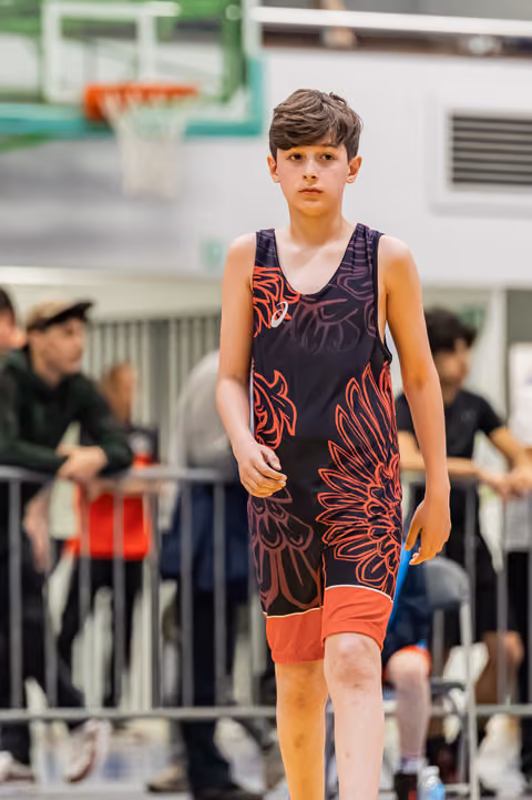 Young boy in ASICS singlet walks with calm focus across gymnasium floor during wrestling event.