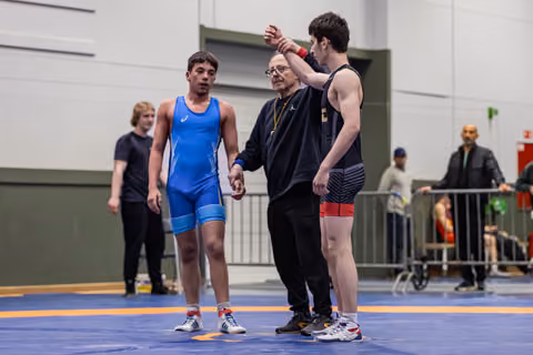 Referee raises the arm of a young wrestler in black, declaring victory, while the defeated opponent in blue stands nearby.