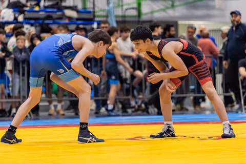 Two young male wrestlers in blue and black singlets face each other in crouched ready stance on yellow mat, crowd behind.