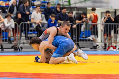 Young wrestler in red singlet dominates opponent in blue, pressing him down on colorful mat before crowd