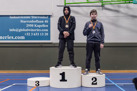 Two young boys stand on a winner's podium; the gold medalist looks composed while the silver medalist proudly displays his medal.