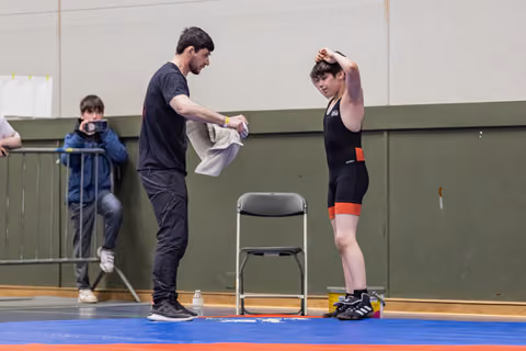 Coach hands a towel to a young wrestler in a black singlet, who stretches arm overhead, resting between bouts on the mat.