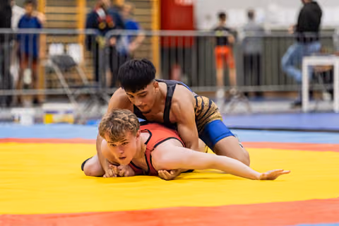 Young wrestler in red singlet pinned to the yellow mat, opponent in black applying pressure in an indoor arena.