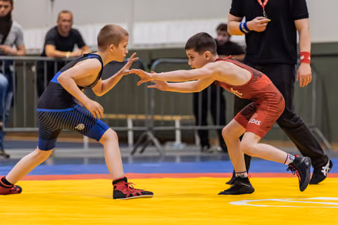 Two young wrestlers face off in focused stances on a yellow mat, arms reaching toward each other during a match.