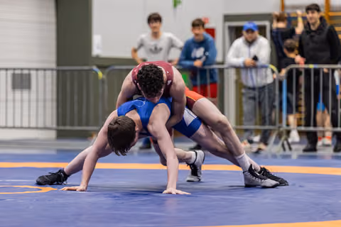 Wrestler in red singlet drives opponent in blue to the mat during an intense youth match indoors.