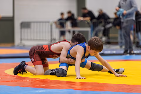 Two young boys wrestle intensely on a colorful mat, one in red controlling the other in blue from behind.