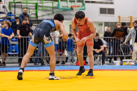 Two wrestlers in blue and red singlets crouch in tense standoff on a yellow mat, crowd watching behind barriers.