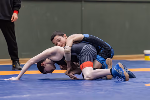 Young wrestler in black singlet controls opponent on blue mat, applying pressure with focused intensity during bout.
