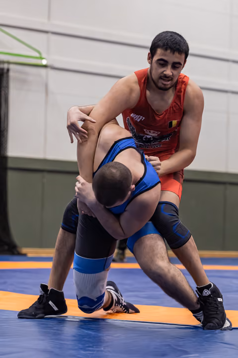 Young wrestler in red singlet dominates opponent in blue, gripping firmly on blue competition mat indoors.