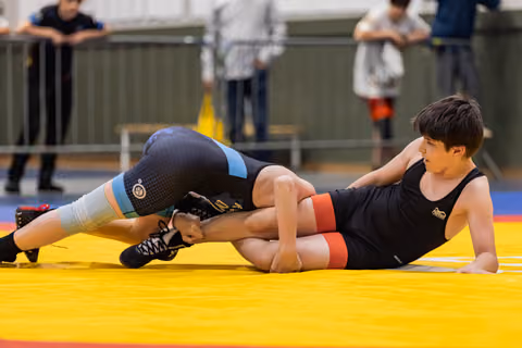 Young boy in black singlet resists leg hold on yellow mat, showing focus and determination during match.
