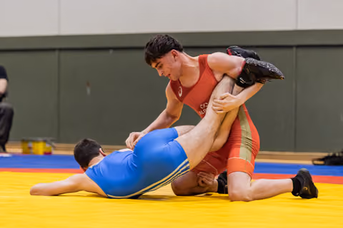 Focused young wrestler in red singlet lifts opponent's leg, dominating blue-clad athlete on yellow mat indoors.