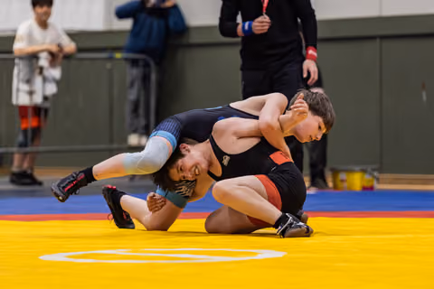 Two young wrestlers battle on a yellow mat, one grimacing as he attempts a takedown during a youth tournament.