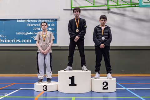 Three young boys stand on a winner's podium, wearing medals, in a sports hall. The first-place boy looks subdued.