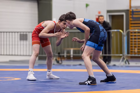Two young wrestlers in red and blue singlets crouch in tense standoff, hands raised, on a competition mat indoors.