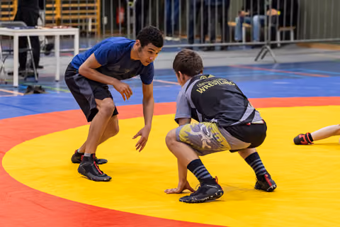 Two young wrestlers in crouched stances face each other intently on a colorful competition mat indoors.
