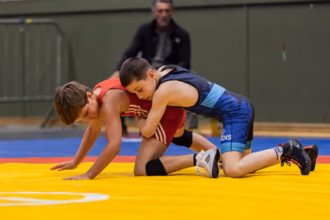 Two young boys wrestle on a yellow mat, one in blue pinning down opponent in red during indoor youth bout.