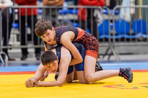 Young wrestler in black-red singlet controls opponent on yellow mat, both focused during intense bout.