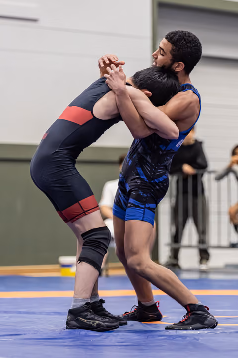 Two young wrestlers locked in a standing clinch on a blue mat, straining with intensity during an indoor match.