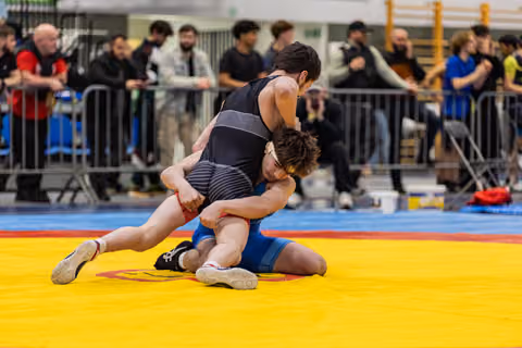 Wrestler in black singlet drives opponent in blue to the mat during an indoor match, crowd watching in background.