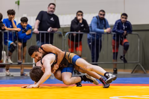 Young wrestler in black singlet drives opponent to the yellow mat while spectators watch intently from behind barriers.