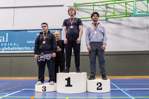 Three teenage wrestlers stand on podiums 1-3, wearing medals, with a coach beside them in an indoor sports hall.