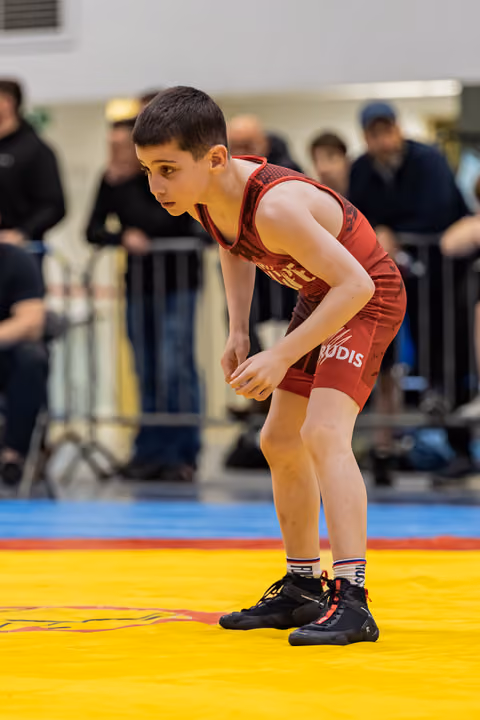 Young boy in red singlet crouches in a focused wrestling stance on a yellow mat, spectators blurred behind.