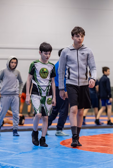 Two young wrestlers walk across the mat, the smaller boy in a green uniform looks downcast, the taller in a Nike hoodie walks beside him.