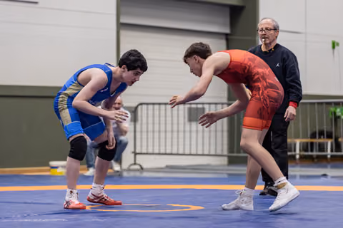 Two young wrestlers in blue and red singlets crouch in tense standoff on blue mat, referee watches closely behind.