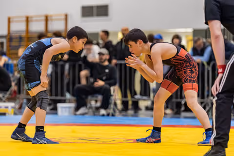 Two young wrestlers in blue and red singlets face off on a yellow mat, both crouched and focused.