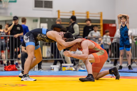 Two wrestlers battle on a yellow mat, one in red kneeling while being controlled in a headlock by opponent in black.