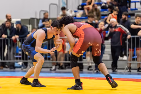 Two young male wrestlers in blue and maroon singlets square off in a crouched stance, crowd watching intently behind barriers.