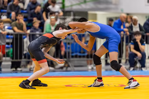 Two young wrestlers in black and blue singlets face off in a tense clinch on a yellow mat before a packed crowd.