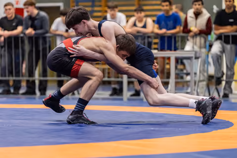 Two young male wrestlers locked in a takedown struggle on a blue and orange mat, spectators watching intently behind barriers.