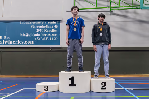 Two boys stand on a podium, first and second place, wearing medals, looking solemn in an indoor sports hall.