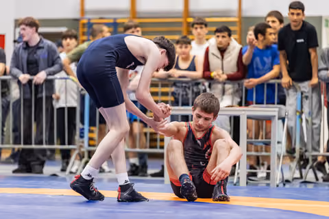 Blue singlet wrestler extends hand to help seated opponent rise from mat, crowd watches in background.