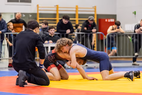 Young wrestler in navy singlet dominates opponent on mat while referee watches closely indoors.