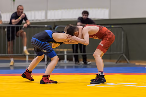 Two young wrestlers lock grips in an intense crouched stance on a yellow mat, watched by spectators behind barriers.