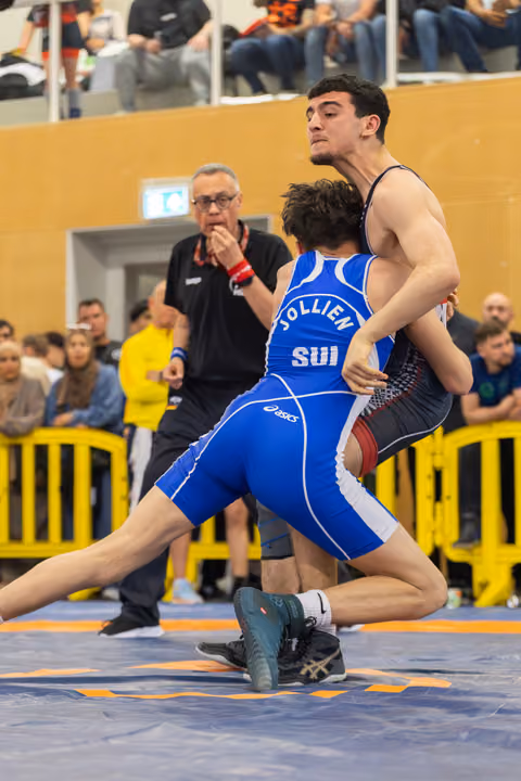 Swiss wrestler Jollien in blue SUI singlet grapples opponent on mat, referee watches intently in background.