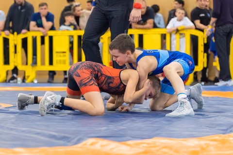 Two young wrestlers compete intensely on a blue mat, one in red dominating over the other in blue during a youth bout.