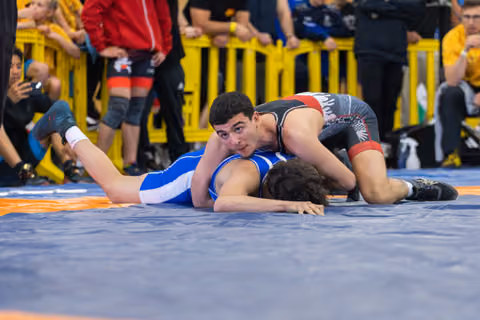 Young wrestler in black singlet pins opponent in blue, looking up confidently during an indoor match.