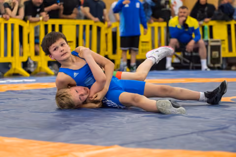 Young boy in blue Adidas singlet pins opponent on mat, looking up with focus during youth wrestling bout.