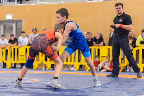 Two boys in red and blue singlets lock grips on the mat, focused and determined, as a referee watches nearby.