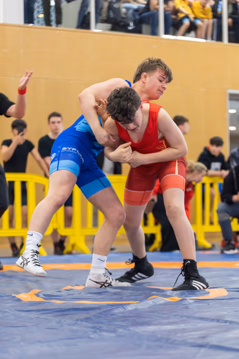 Two young wrestlers grapple fiercely on a blue mat, one in blue lunging forward, the other in red bracing defensively.